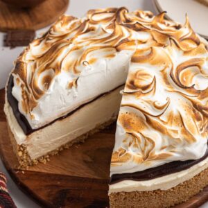 A No Bake S'mores Cheesecake with toasted meringue topping sits on a wooden board, one slice missing.