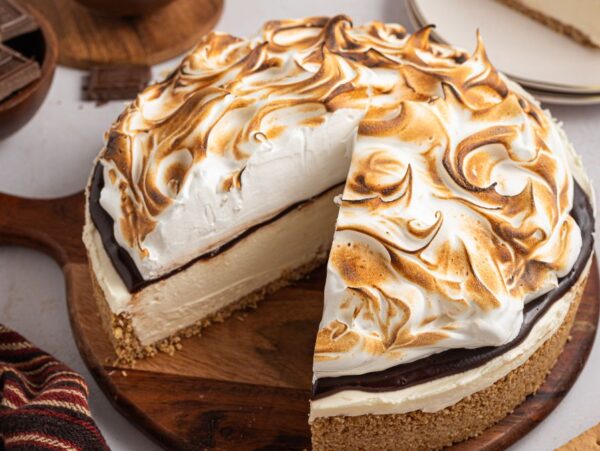 A No Bake S'mores Cheesecake with toasted meringue topping sits on a wooden board, one slice missing.