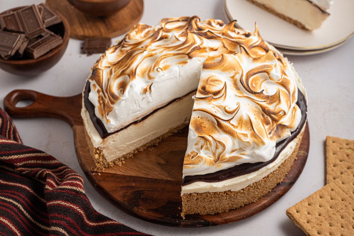 A No Bake S'mores Cheesecake with toasted meringue topping sits on a wooden board, one slice cut, surrounded by graham crackers.