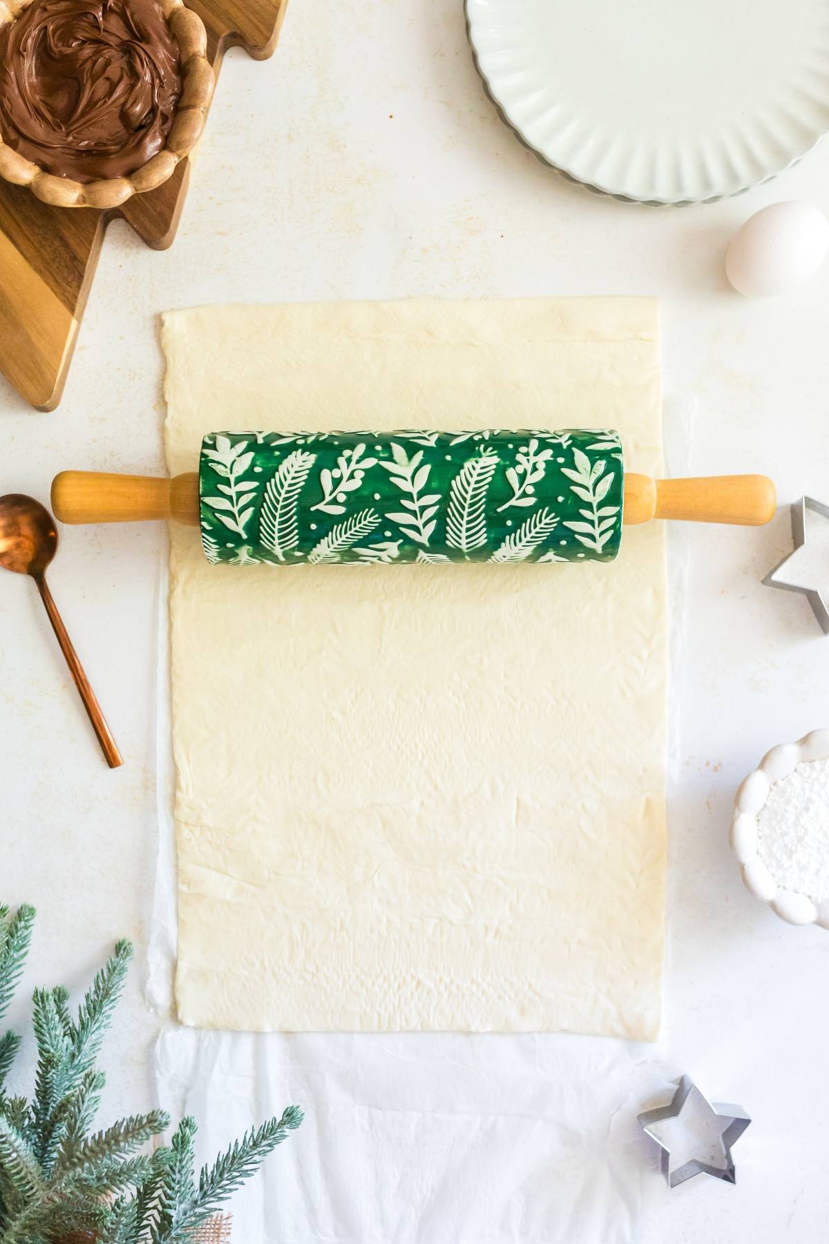 A green rolling pin on puff pastry, surrounded by baking tools, an egg, and a sprig of pine.