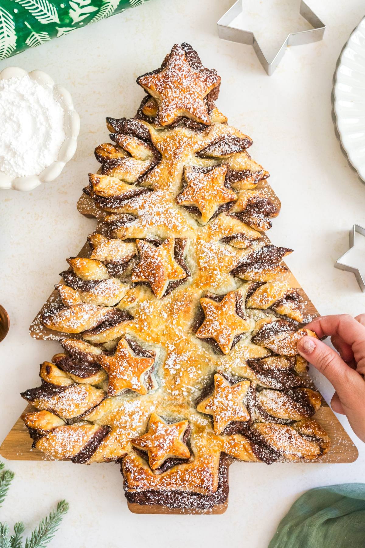Tree-shaped pastry with star cutouts, dusted with powdered sugar. Hand reaches for a piece.