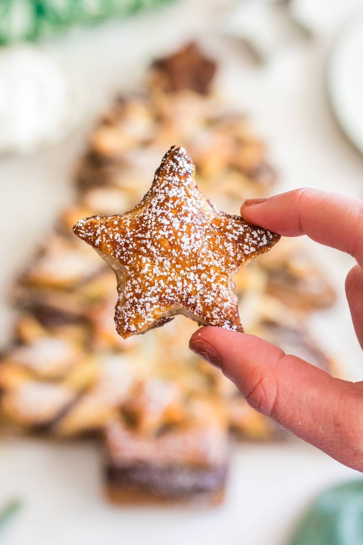 A hand holds a star-shaped pastry dusted with powdered sugar, with a blurred Christmas tree pastry in the background.