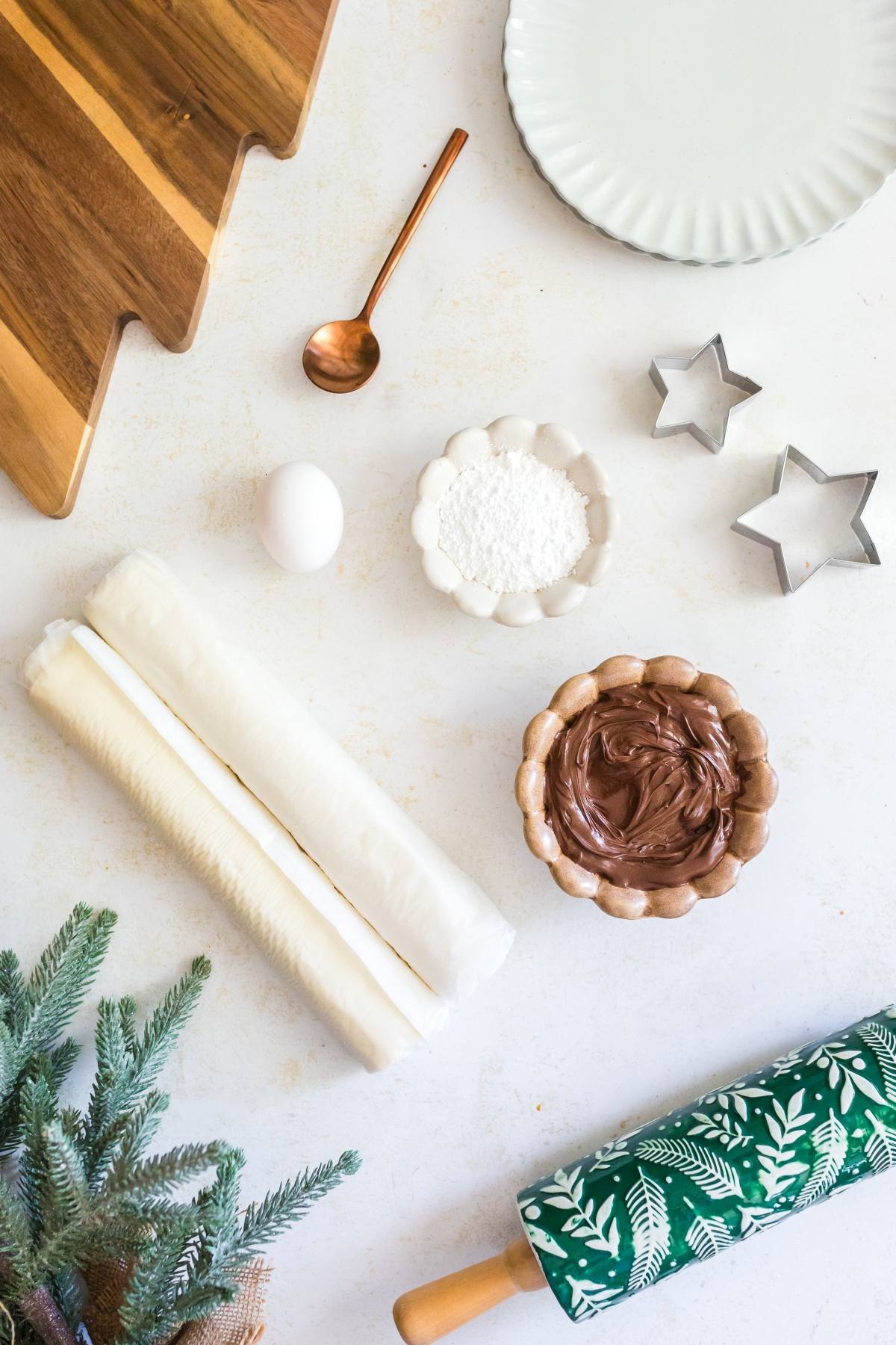 Baking ingredients and tools on a white surface, including dough, egg, Nutella, powdered sugar, and cookie cutters.