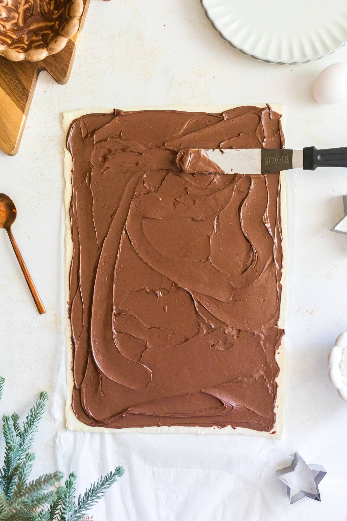 A spatula spreads chocolate filling on a rectangular sheet of pastry dough on a white surface.