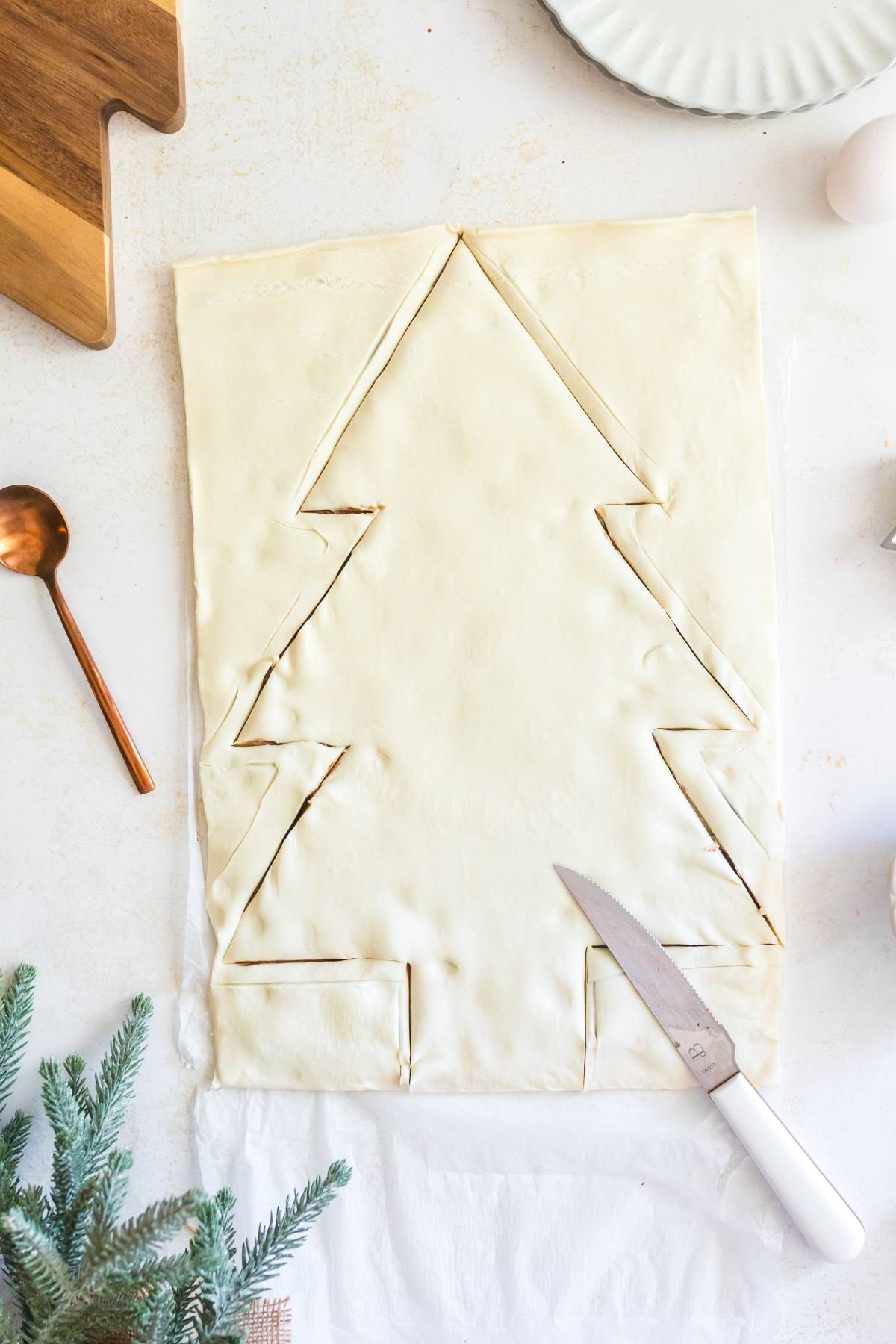Rectangle of pastry dough with a Christmas tree shape cut out, knife and kitchen items nearby on white surface.
