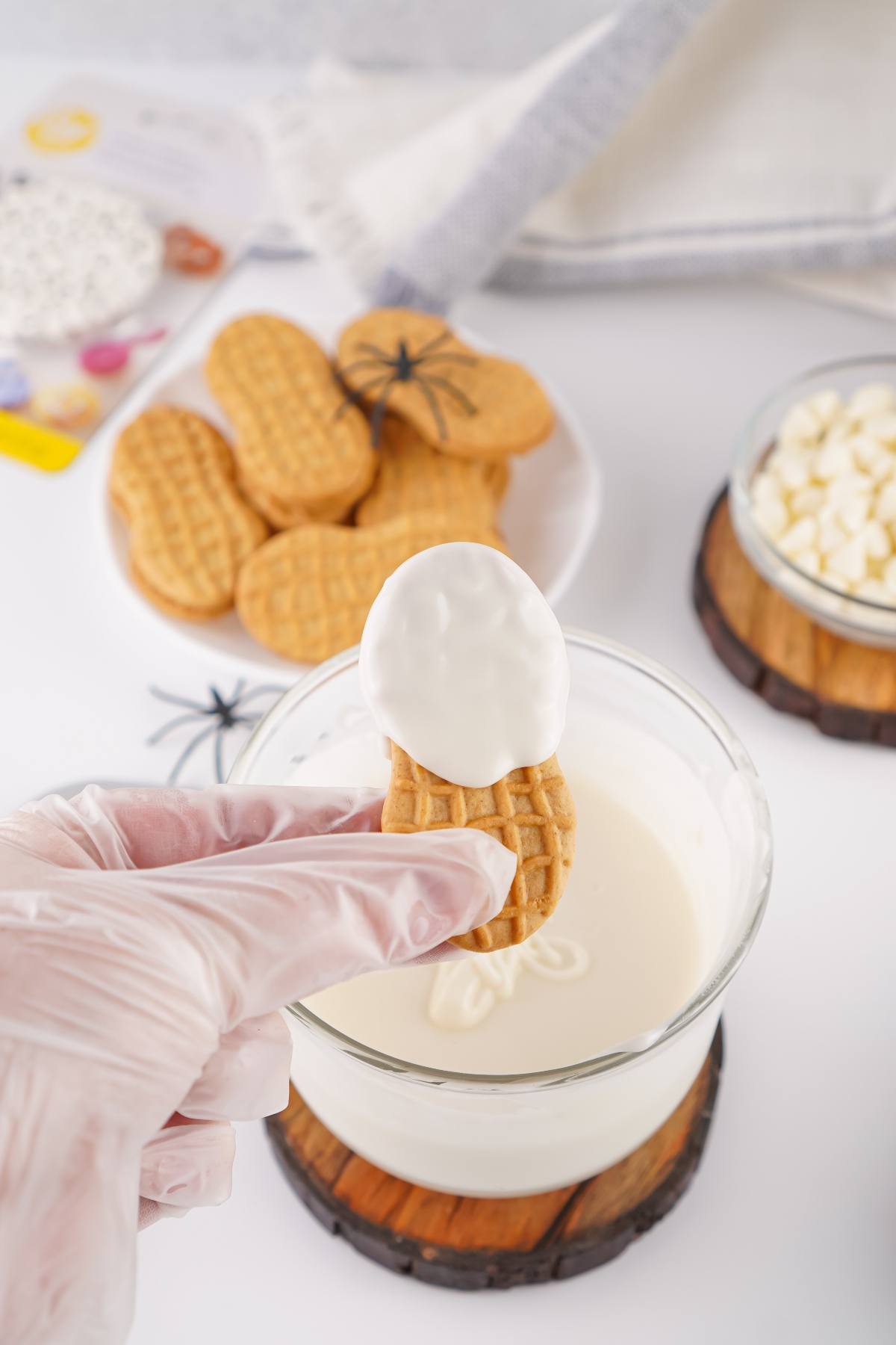 A gloved hand dips a peanut-shaped cookie into melted white chocolate, with cookies and chocolate nearby.