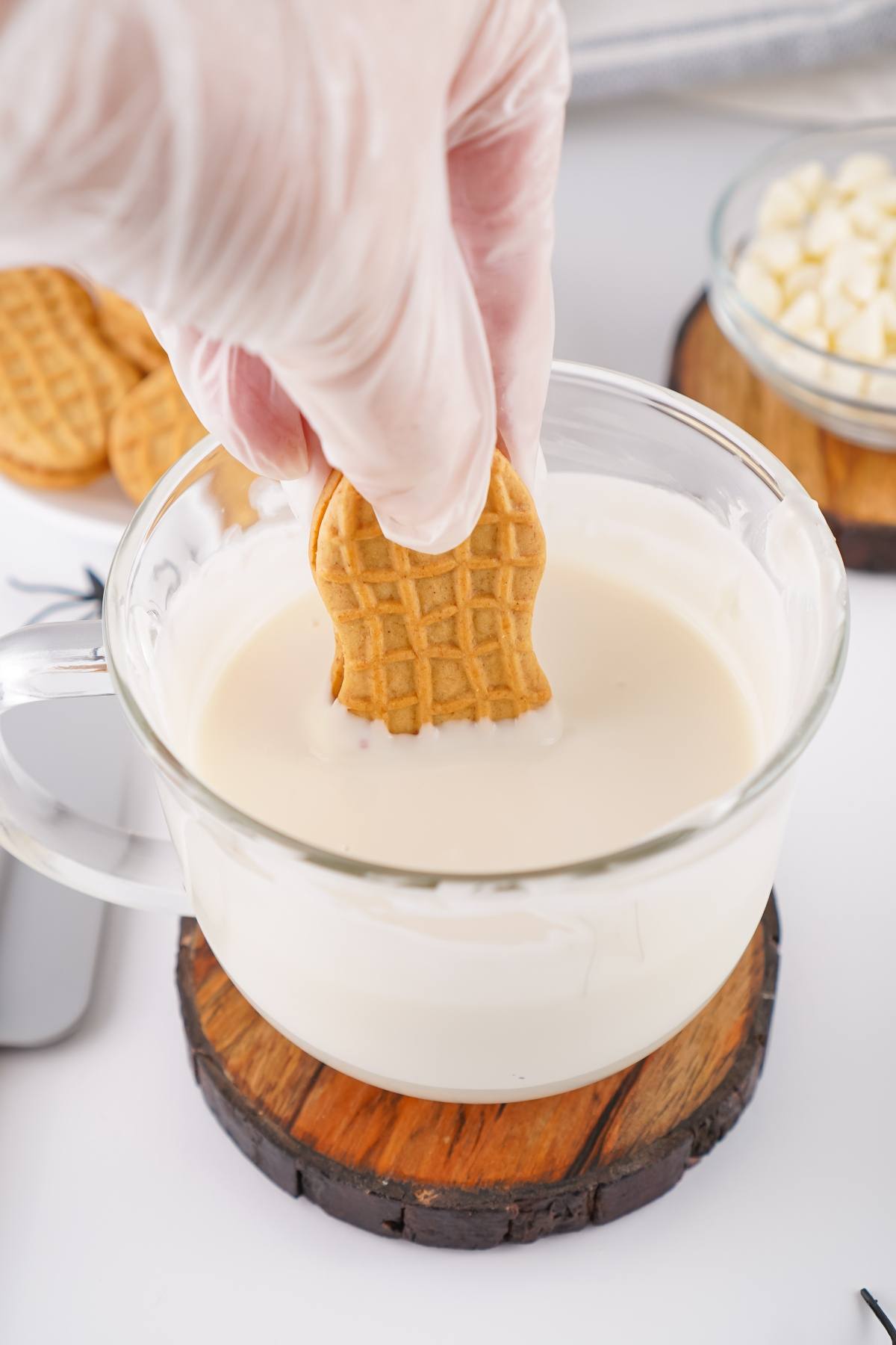 A gloved hand dips a peanut butter cookie into a glass cup of melted white chocolate.
