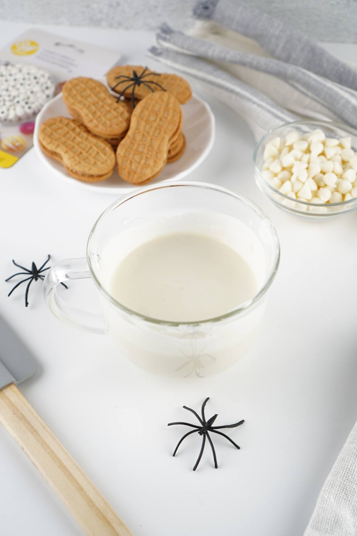 A glass cup of milk with cookies, white chocolate chips, and fake black spiders on a white table.