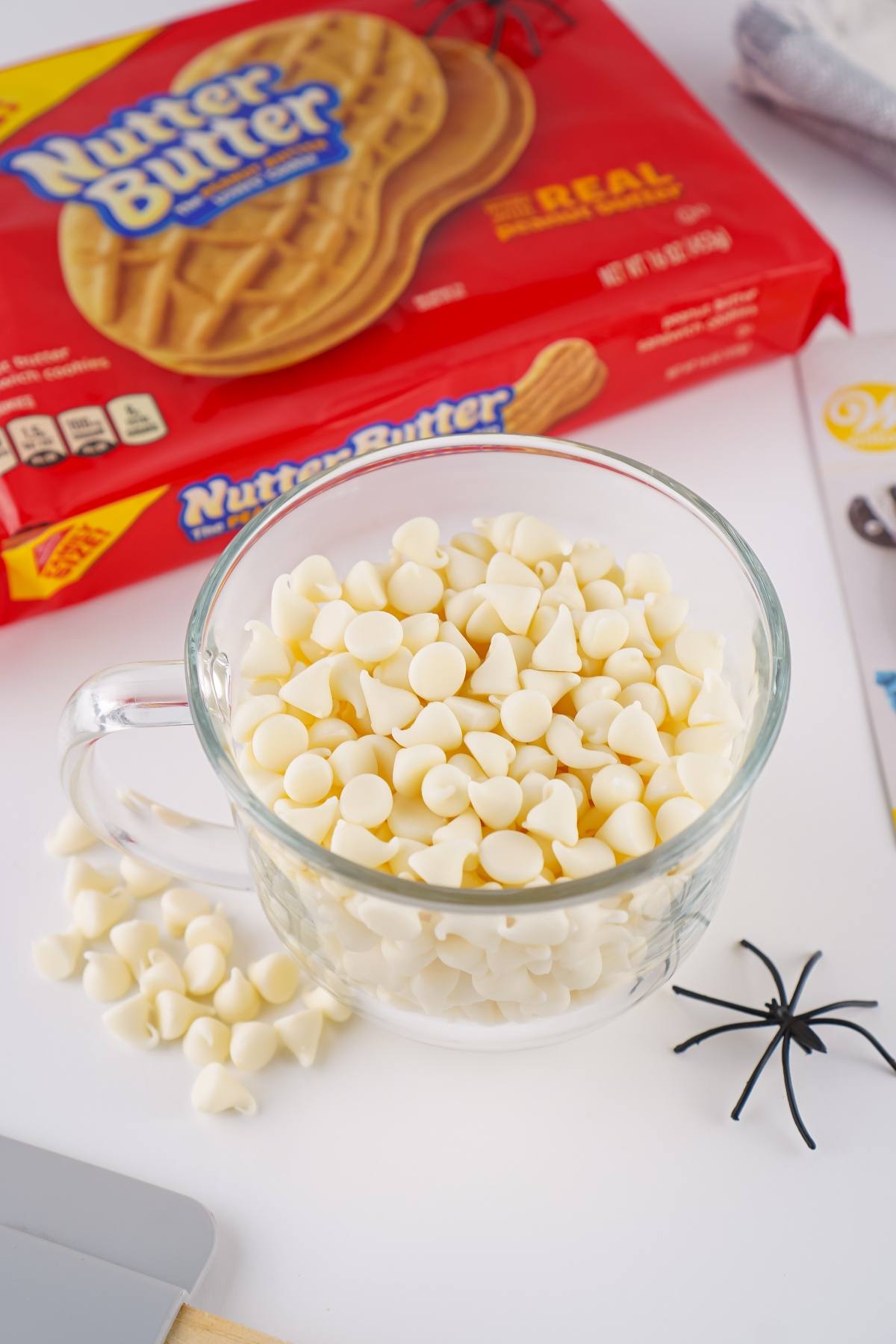 A glass cup filled with white chocolate chips sits near Nutter Butter cookies and a plastic black spider.