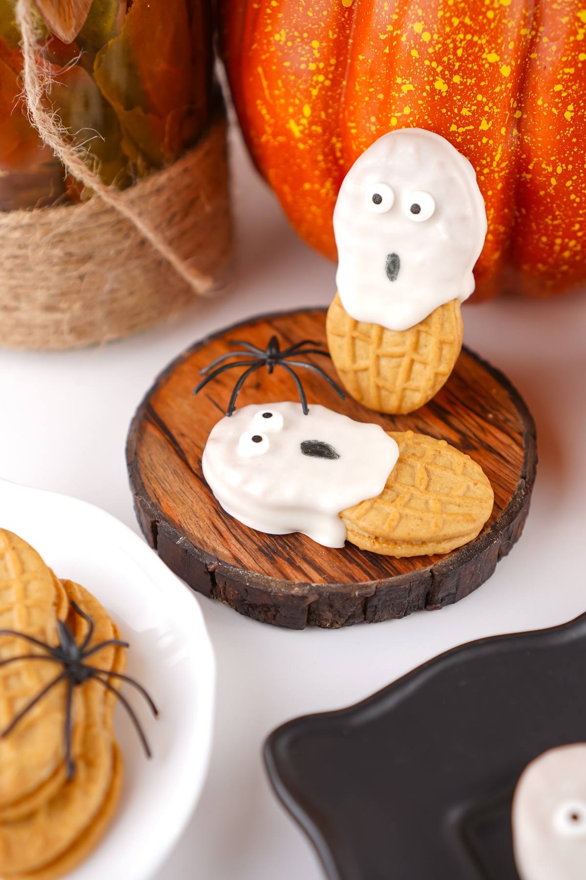 Two peanut-shaped cookies decorated as white chocolate ghosts with candy eyes, next to fake spiders and pumpkins.