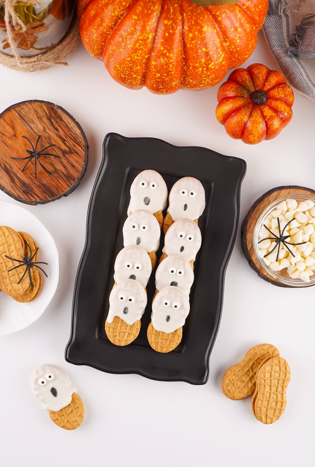 Peanut butter cookies decorated as white ghosts on a black tray, surrounded by pumpkins and fake spiders.