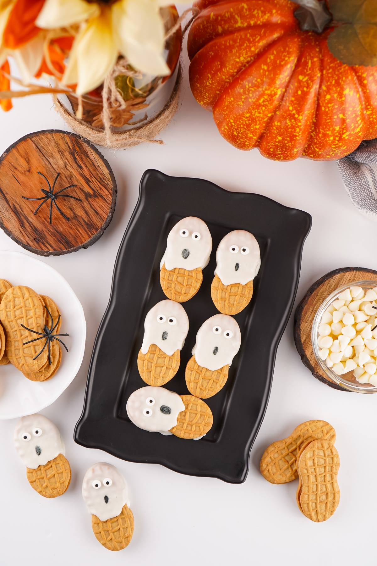 Peanut cookies decorated as white chocolate ghosts on a black tray, surrounded by fall decor and pumpkin.