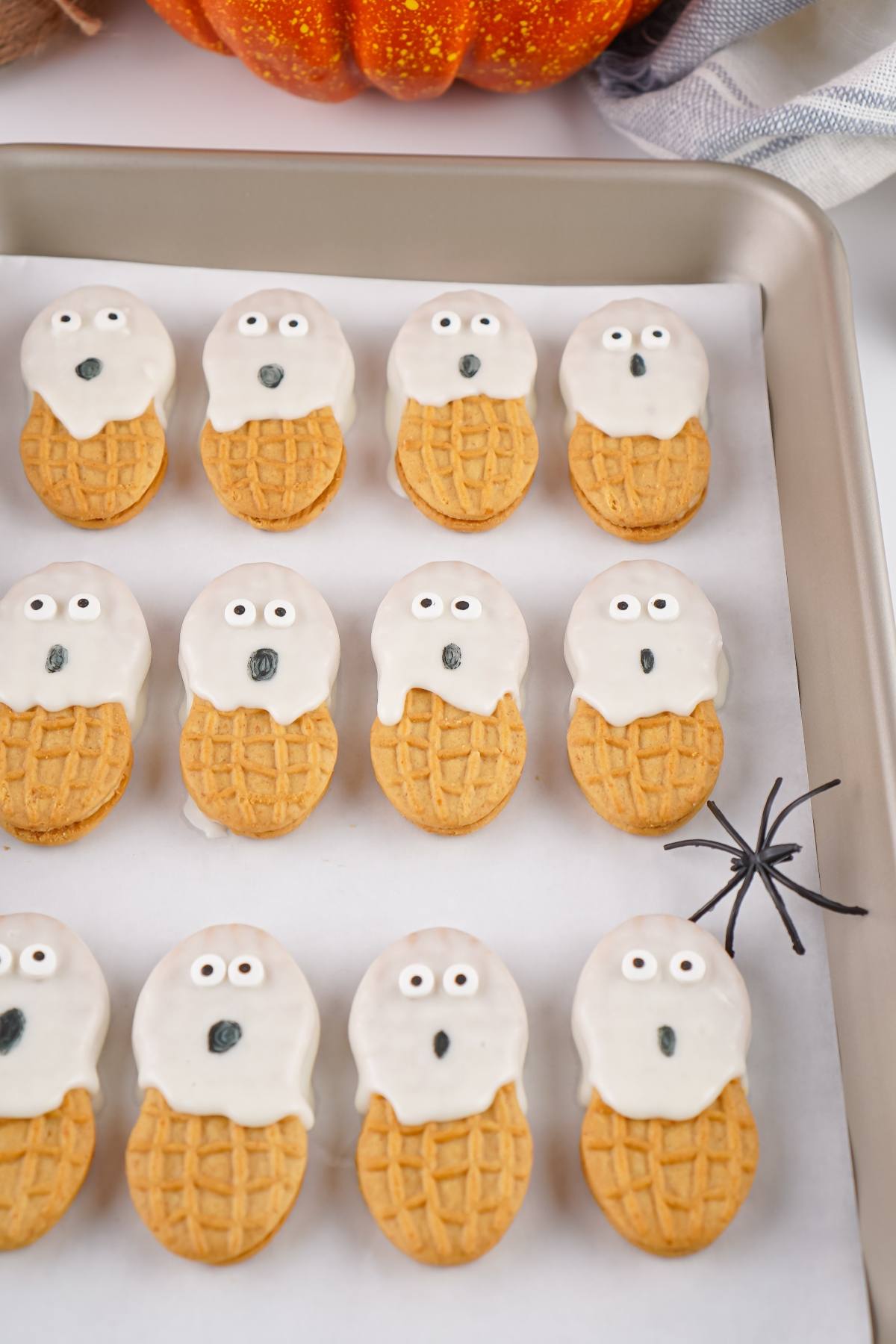 Nutter Butter cookies decorated as ghosts with white icing and candy eyes on a baking tray, with a fake spider nearby.