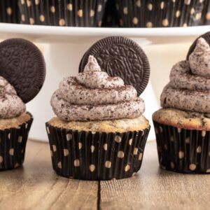 Three Oreo cupcakes with cookies and cream frosting and an Oreo on top, in black polka dot wrappers, sit on a wooden table.