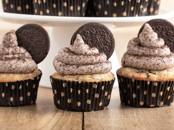 Three Oreo cupcakes with cookies and cream frosting and an Oreo on top, in black polka dot wrappers, sit on a wooden table.