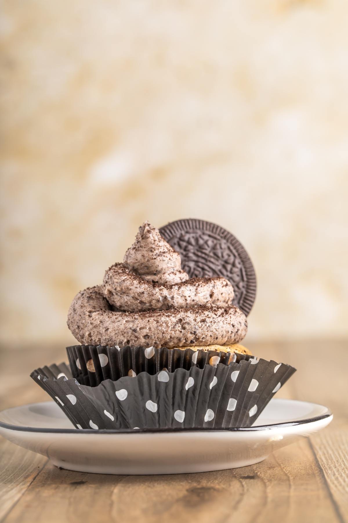 Cookies and cream cupcake with swirled frosting and an Oreo cookie on top, on a plate with a polka dot wrapper.