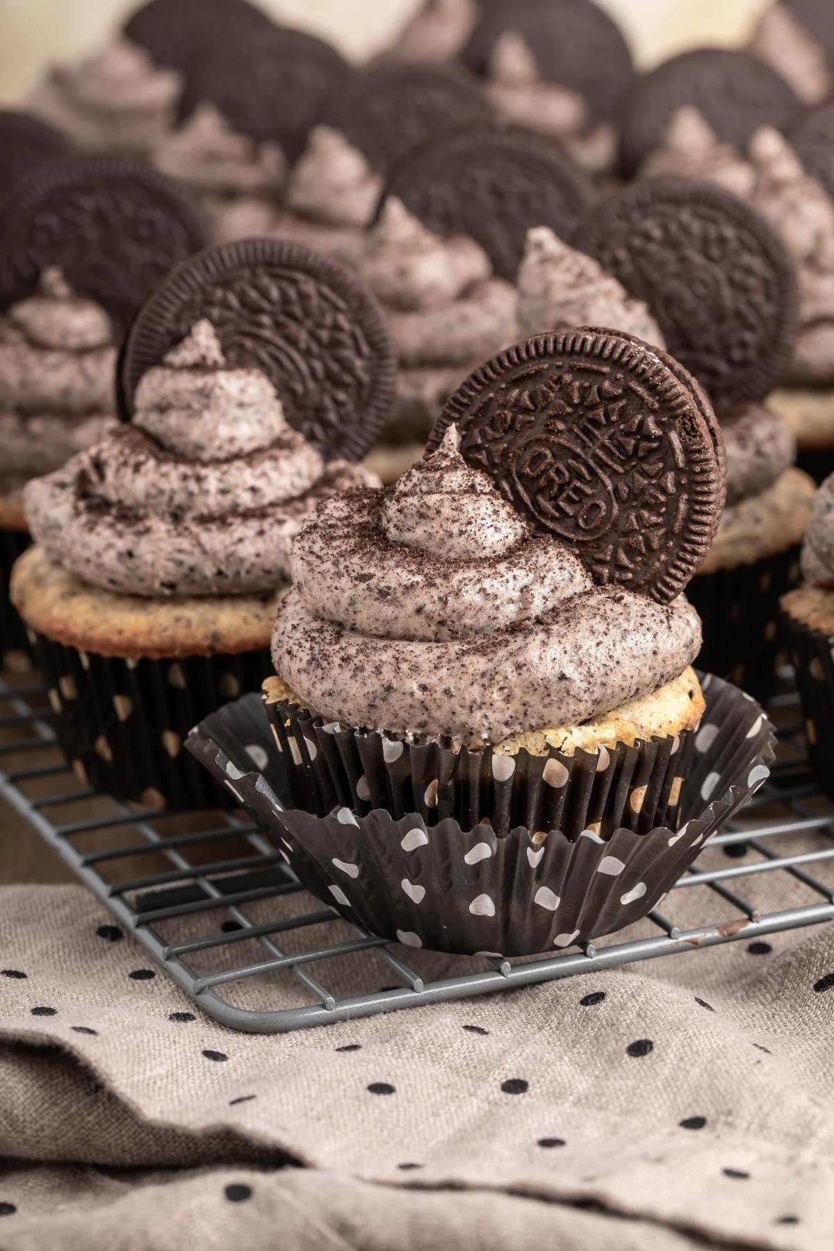 Cookies and cream cupcakes with Oreo cookies on top, displayed on a cooling rack and a spotted cloth.