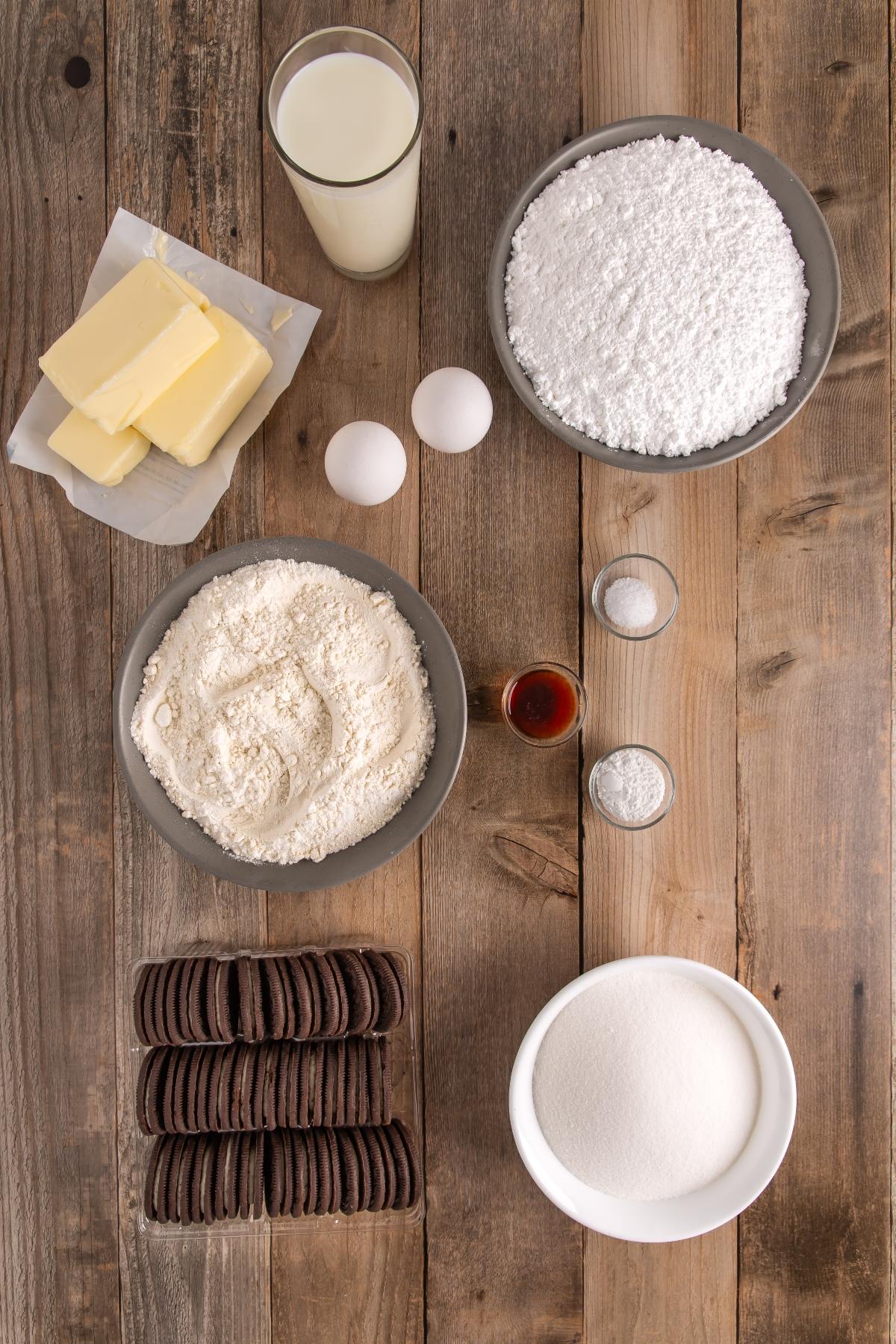 Baking ingredients on a wooden table: butter, eggs, milk, flour, sugar, cookies, vanilla, and powders in bowls.