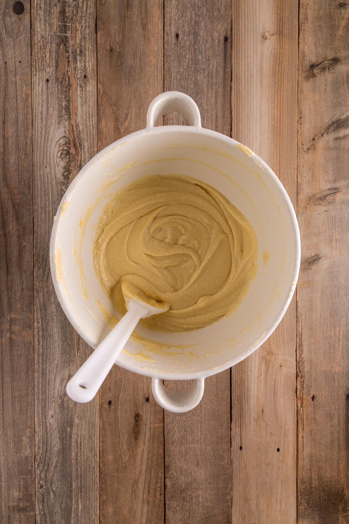A white mixing bowl with light brown batter and a white spatula on a wooden surface.