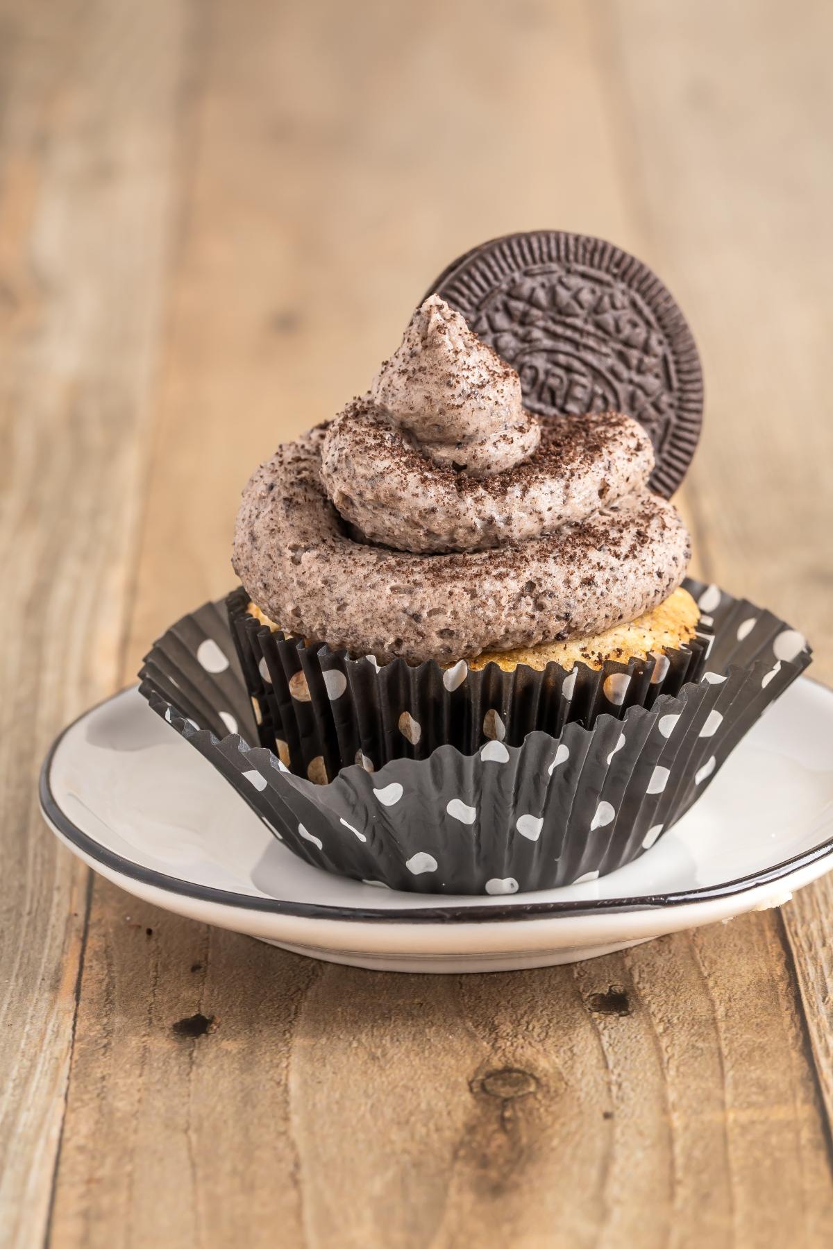 Cupcake with cookies and cream frosting and a chocolate cookie on top, in a polka dot wrapper on a plate.