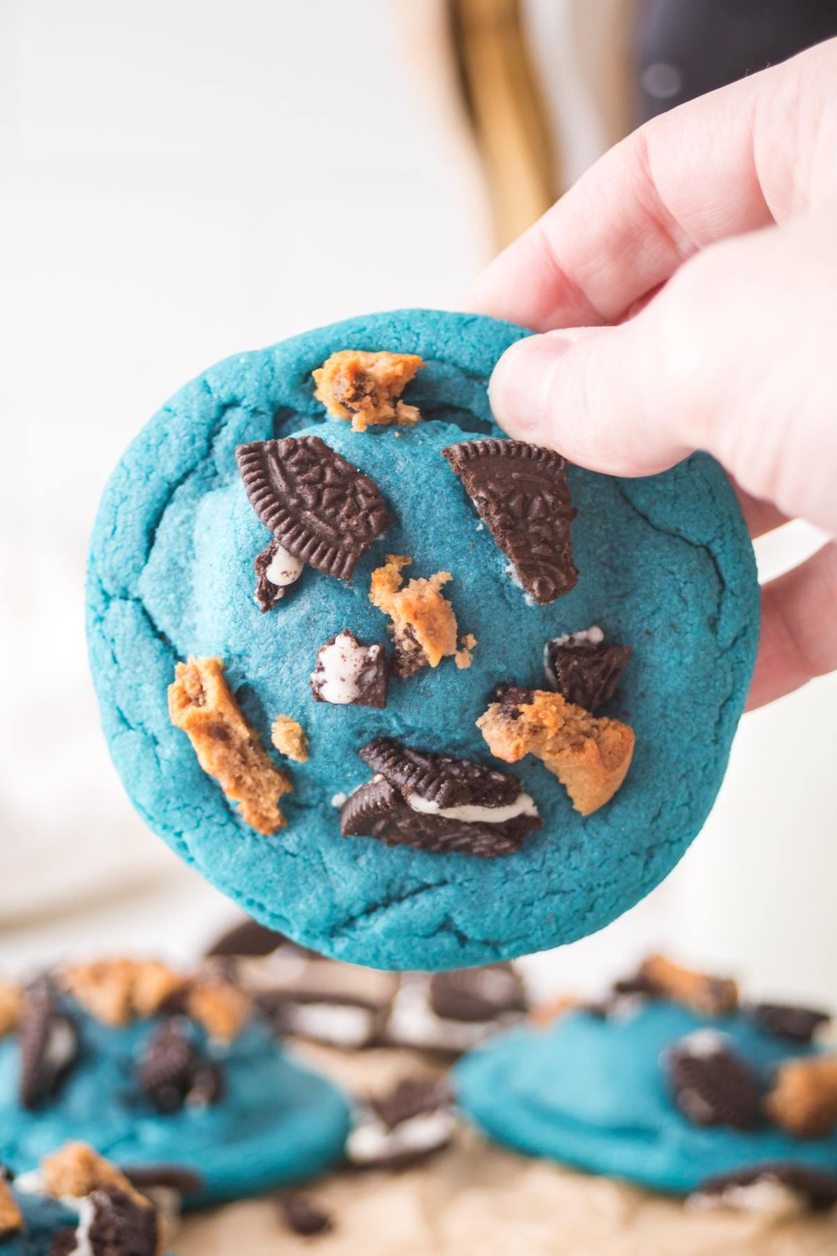 A hand holding a bright blue cookie topped with Oreo pieces and cookie chunks.