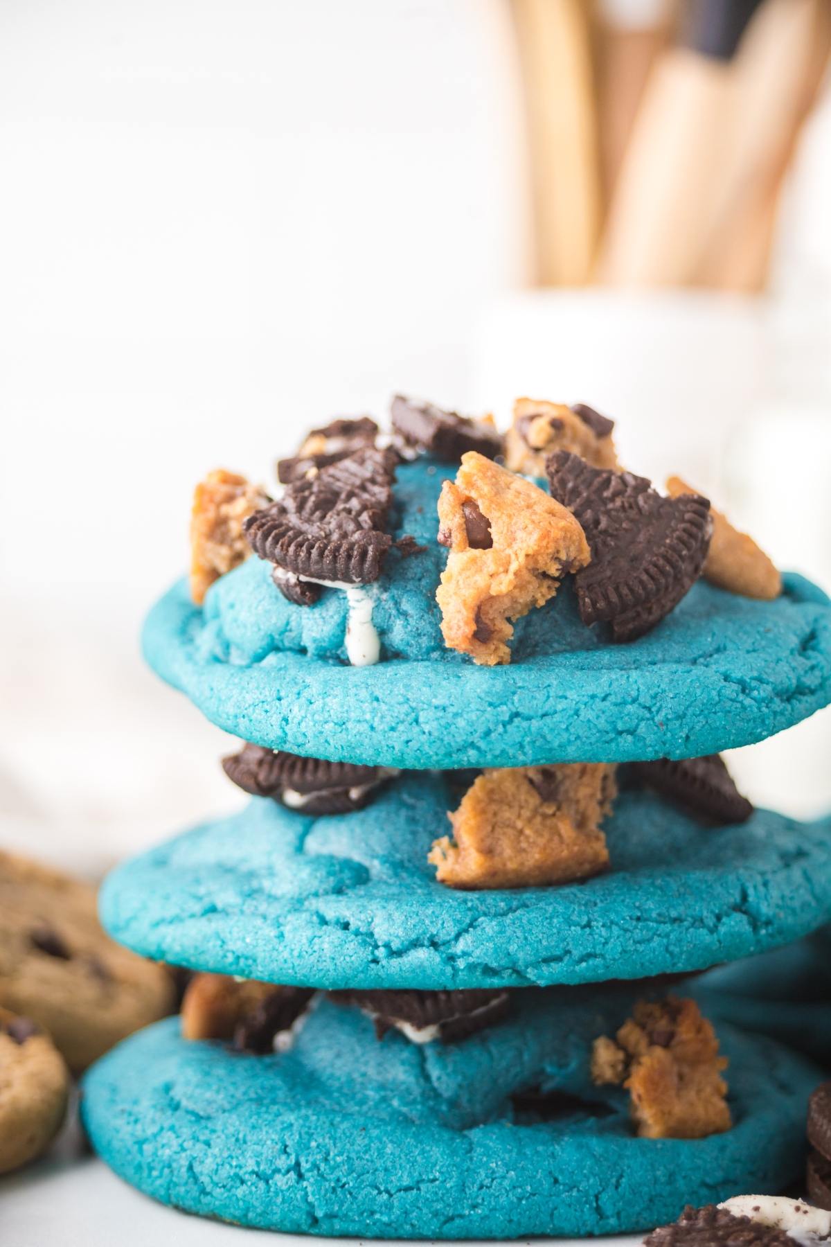 Three stacked blue cookies topped with cookie and chocolate pieces, with blurred kitchen utensils in the background.