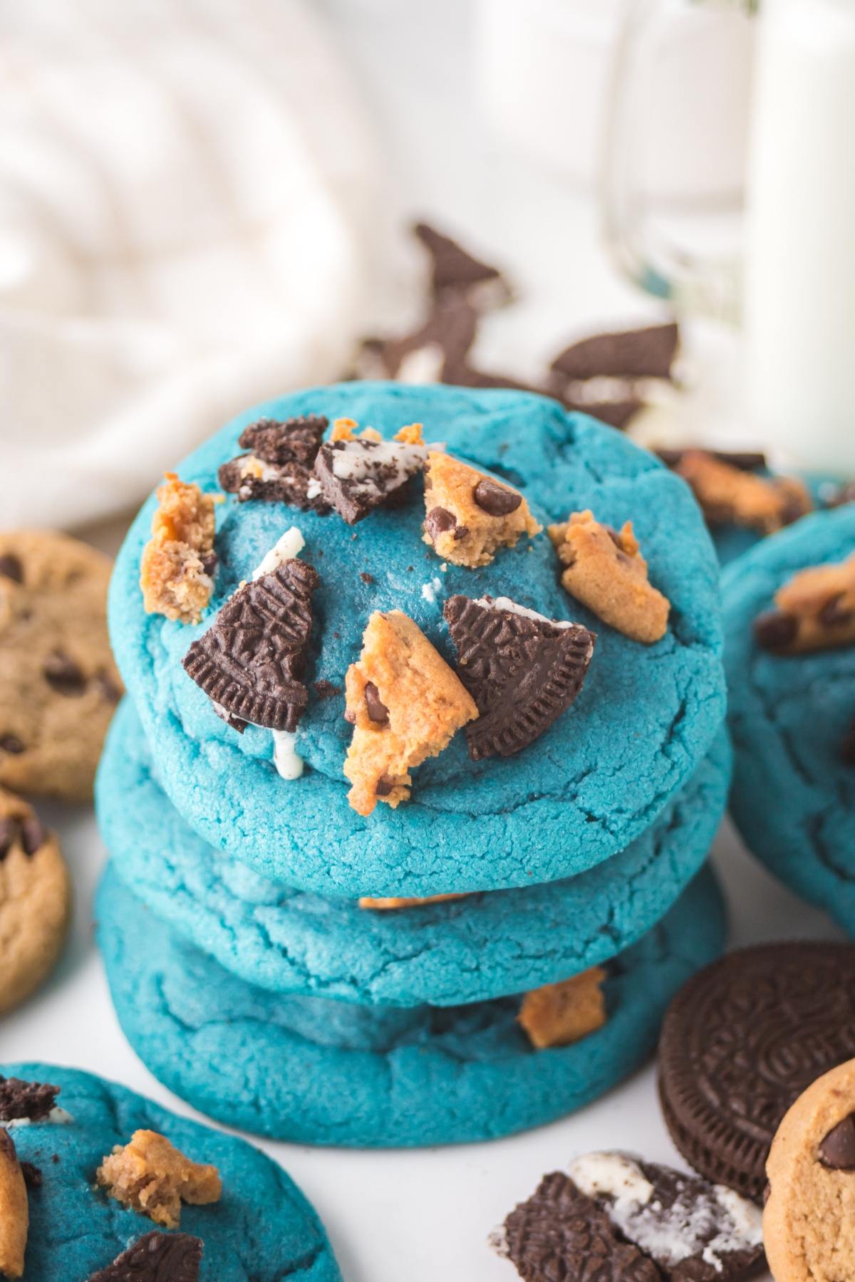 Stack of blue cookies topped with chocolate chip cookie and Oreo pieces, with more cookies around.