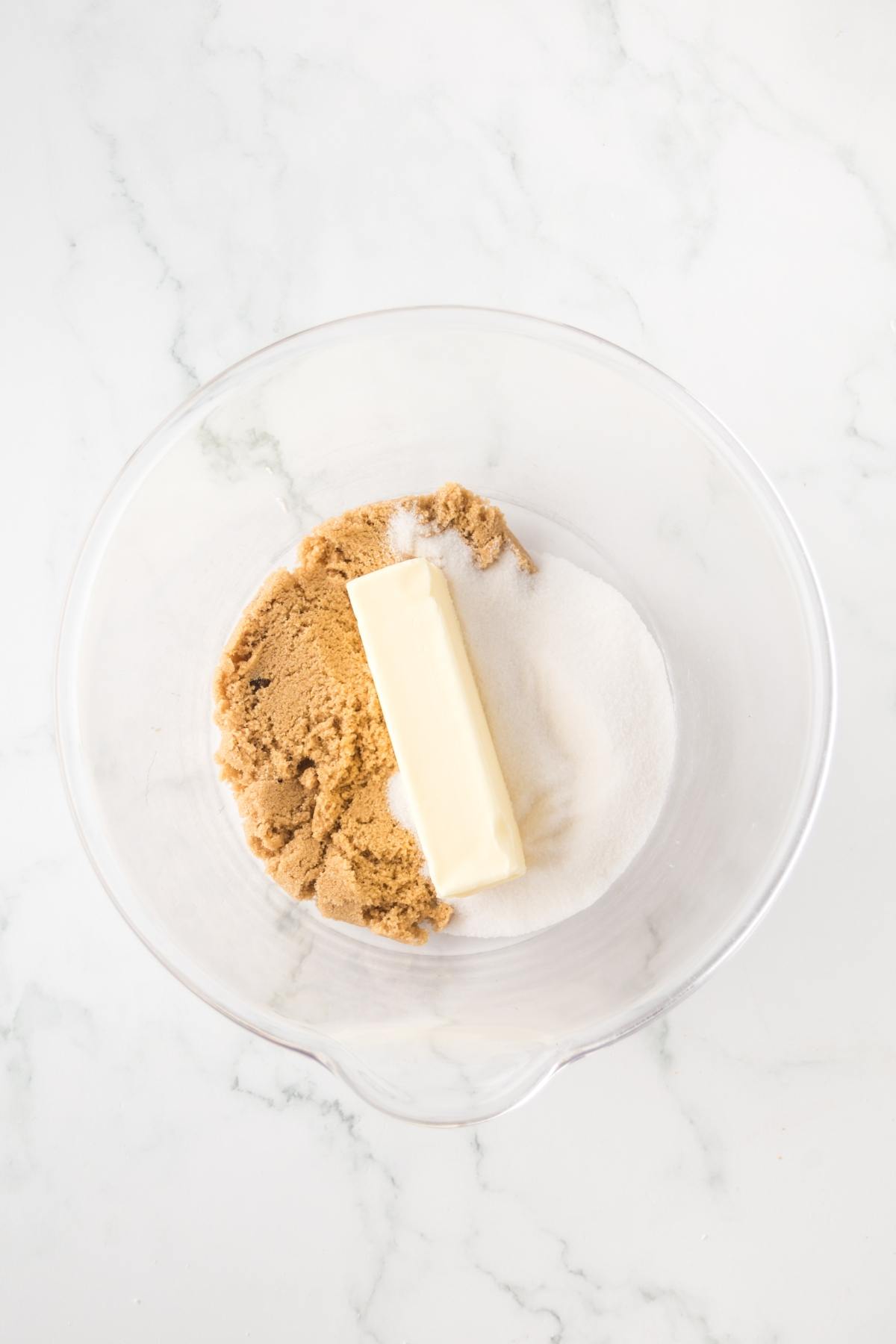 A glass bowl with brown sugar, white sugar, and a stick of butter on a white marble surface.