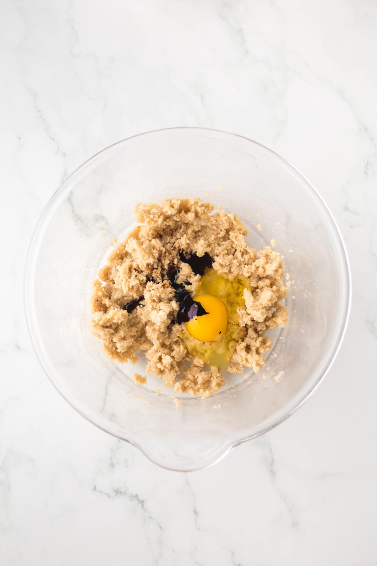 A glass bowl with cookie dough, an egg, and vanilla extract on a white marble surface.