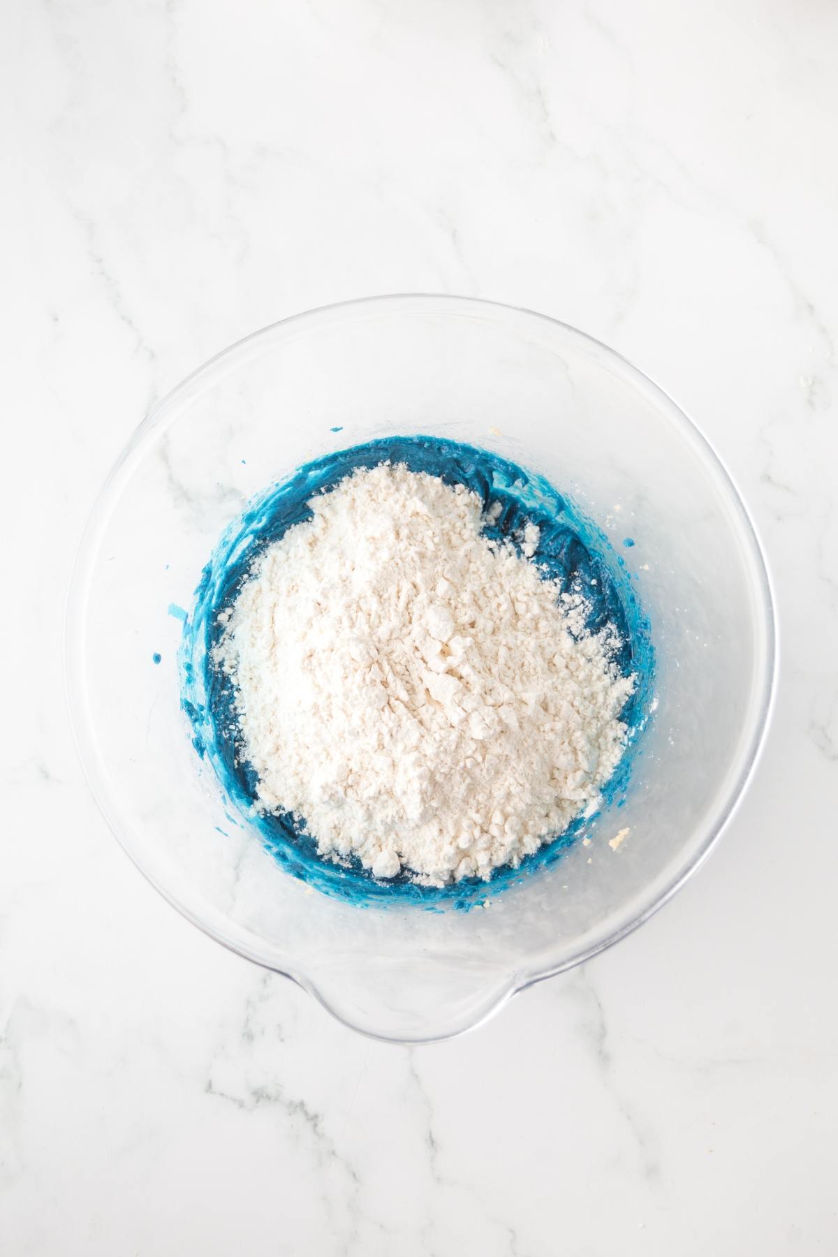 A glass mixing bowl with blue dough and white flour on a white marble surface.