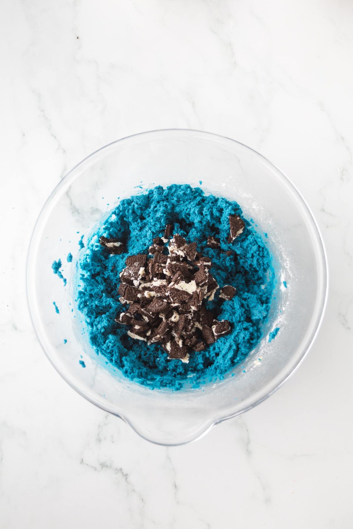 Clear bowl with bright blue cookie dough and crumbled chocolate sandwich cookies on top, on a white surface.