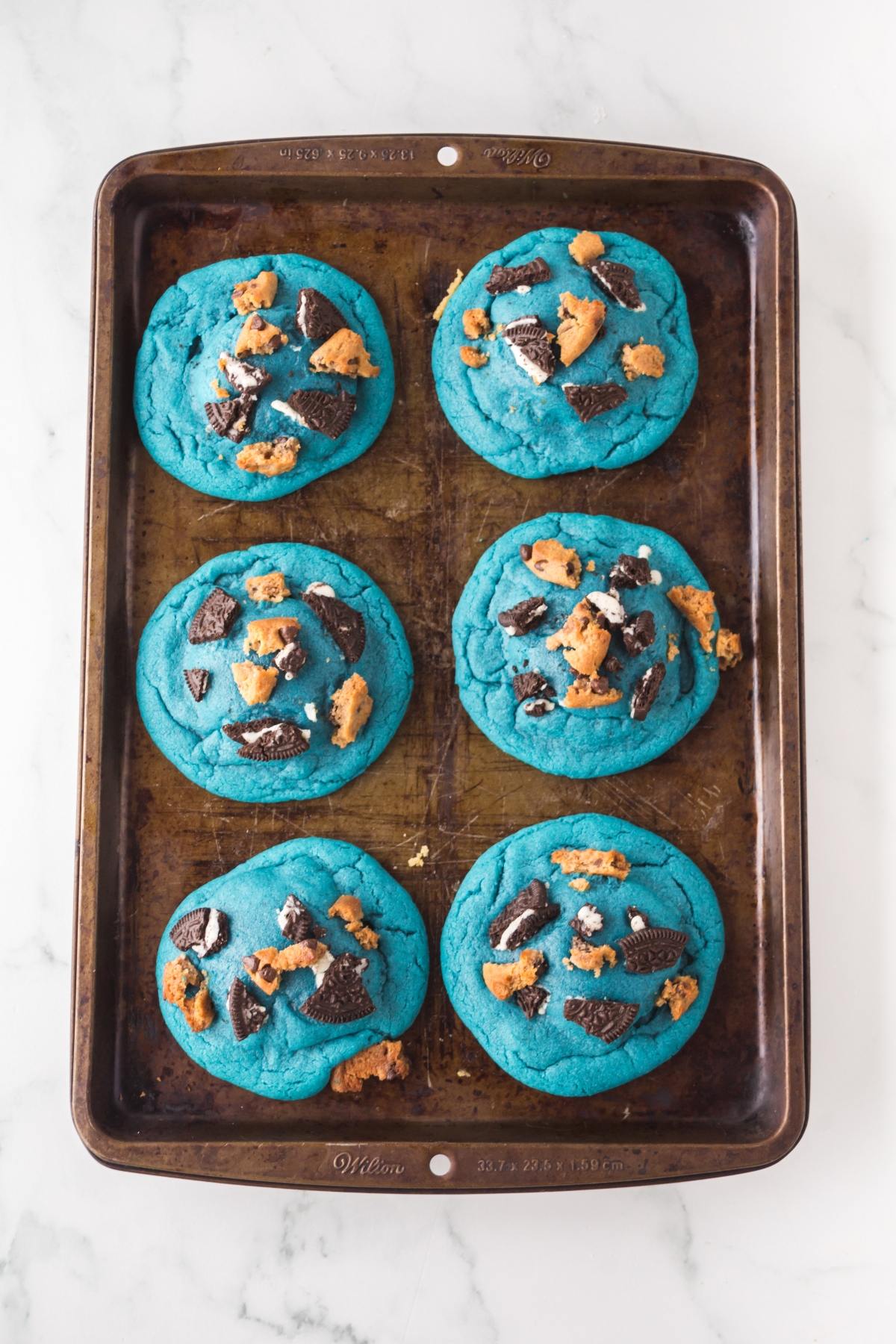Six large blue cookies with cookie and chocolate pieces on a baking sheet, viewed from above.