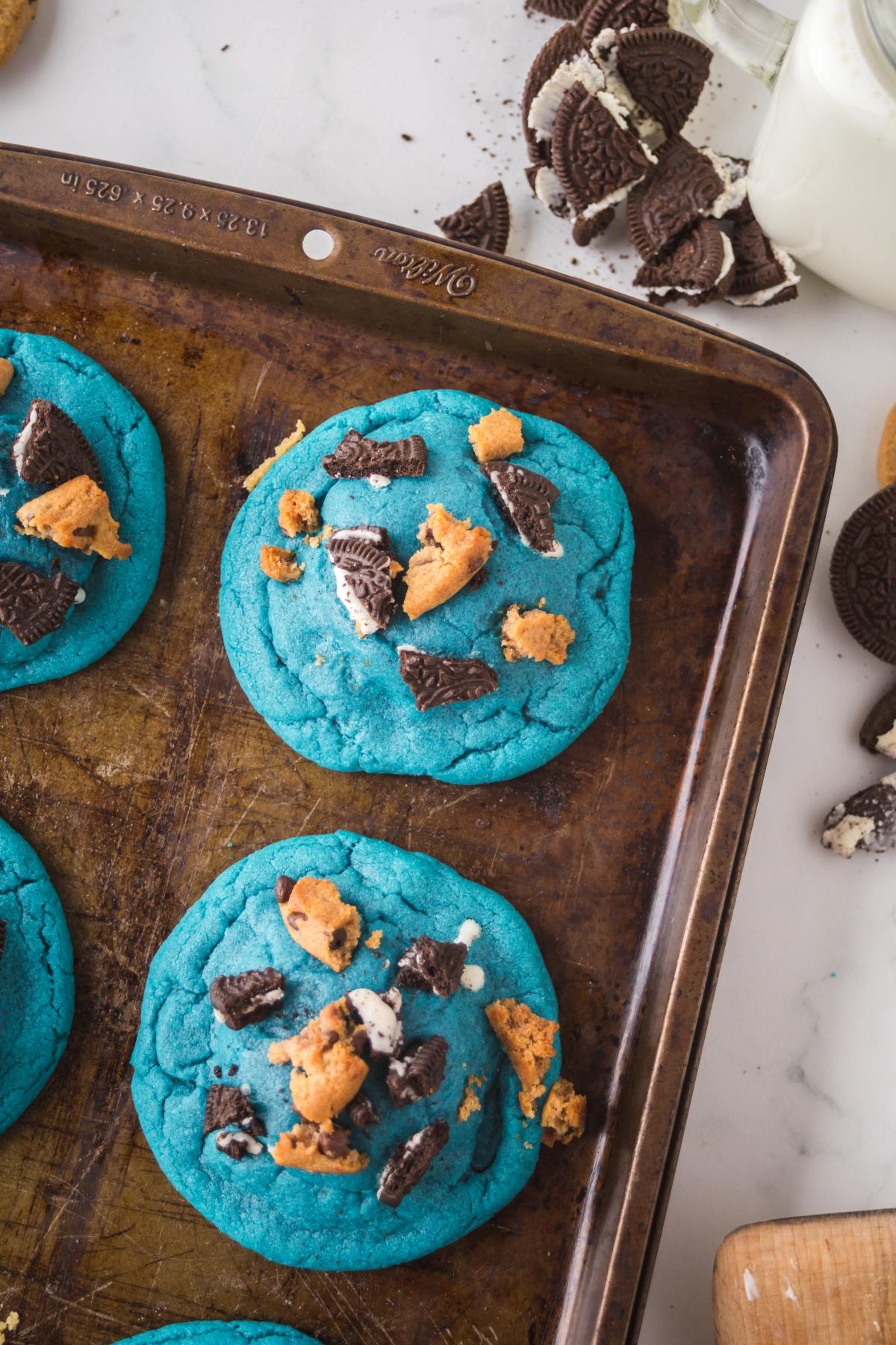 Blue cookies topped with crushed chocolate and vanilla sandwich cookies on a baking tray, with milk nearby.