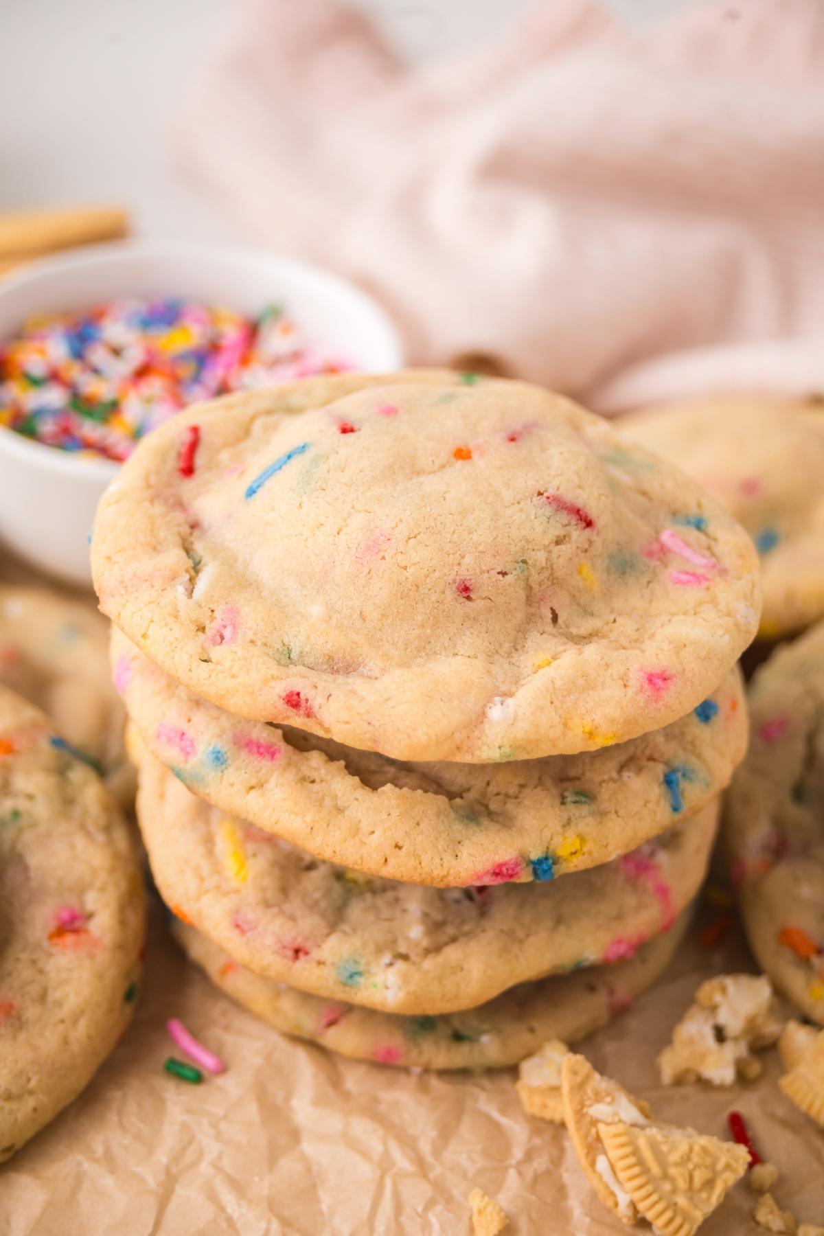 A stack of funfetti cookies with colorful sprinkles, with more cookies and a bowl of sprinkles in the background.