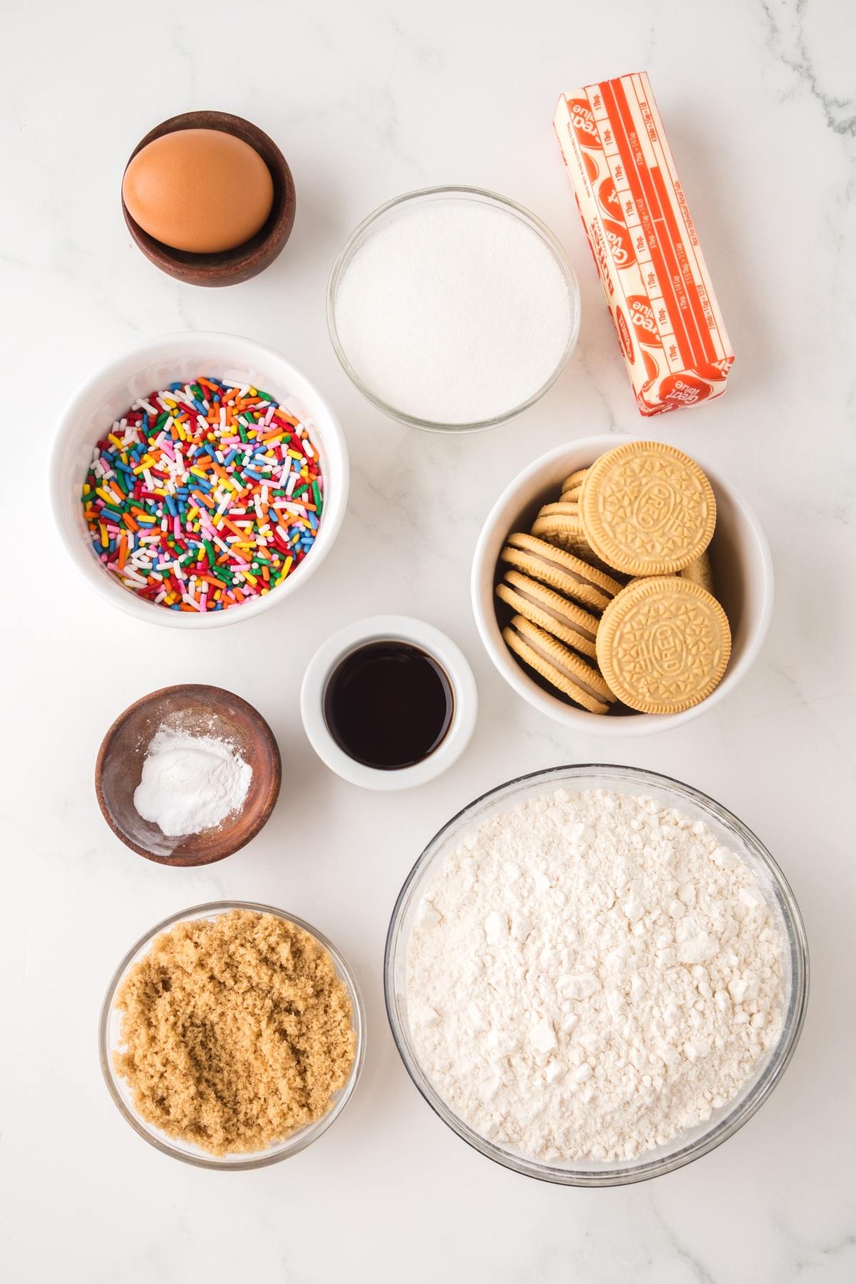Baking ingredients: egg, sugar, butter, sprinkles, vanilla, cookies, flour, brown sugar, baking powder on white surface.