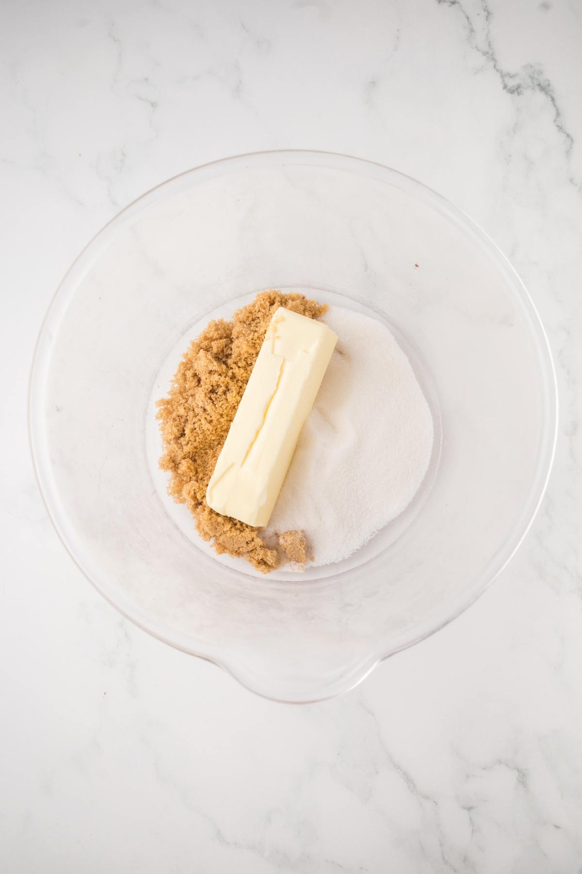 A glass bowl with butter, brown sugar, and white sugar on a marble surface.