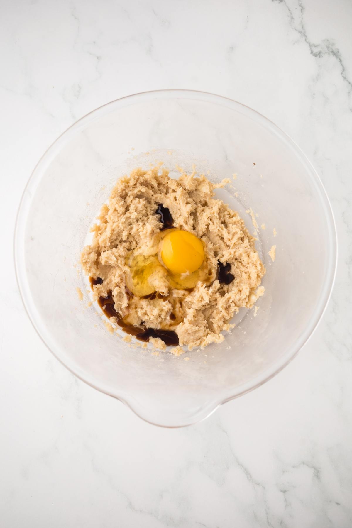 A mixing bowl with cookie dough, an egg, and vanilla extract on a white marble surface.