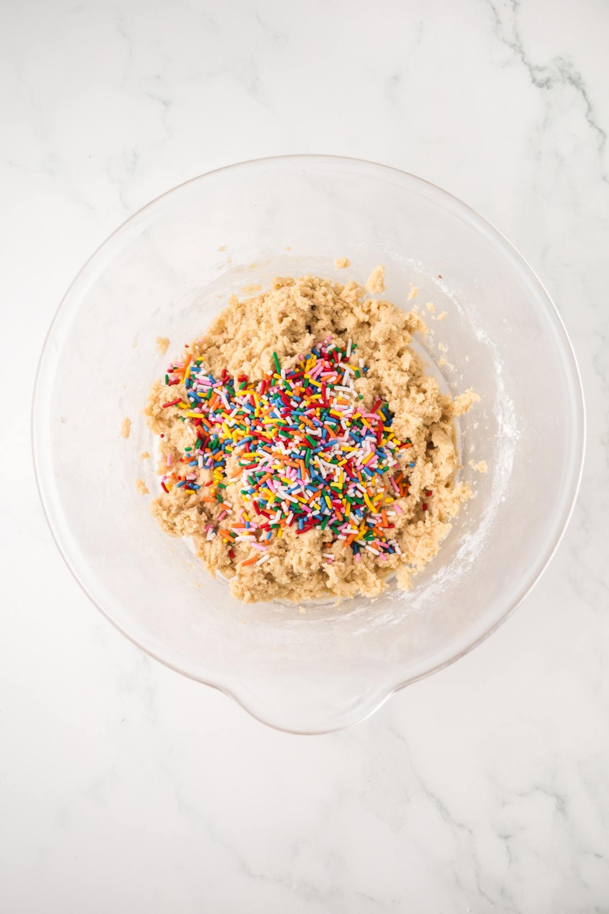 Clear glass bowl with cookie dough and colorful sprinkles on a white marble countertop.
