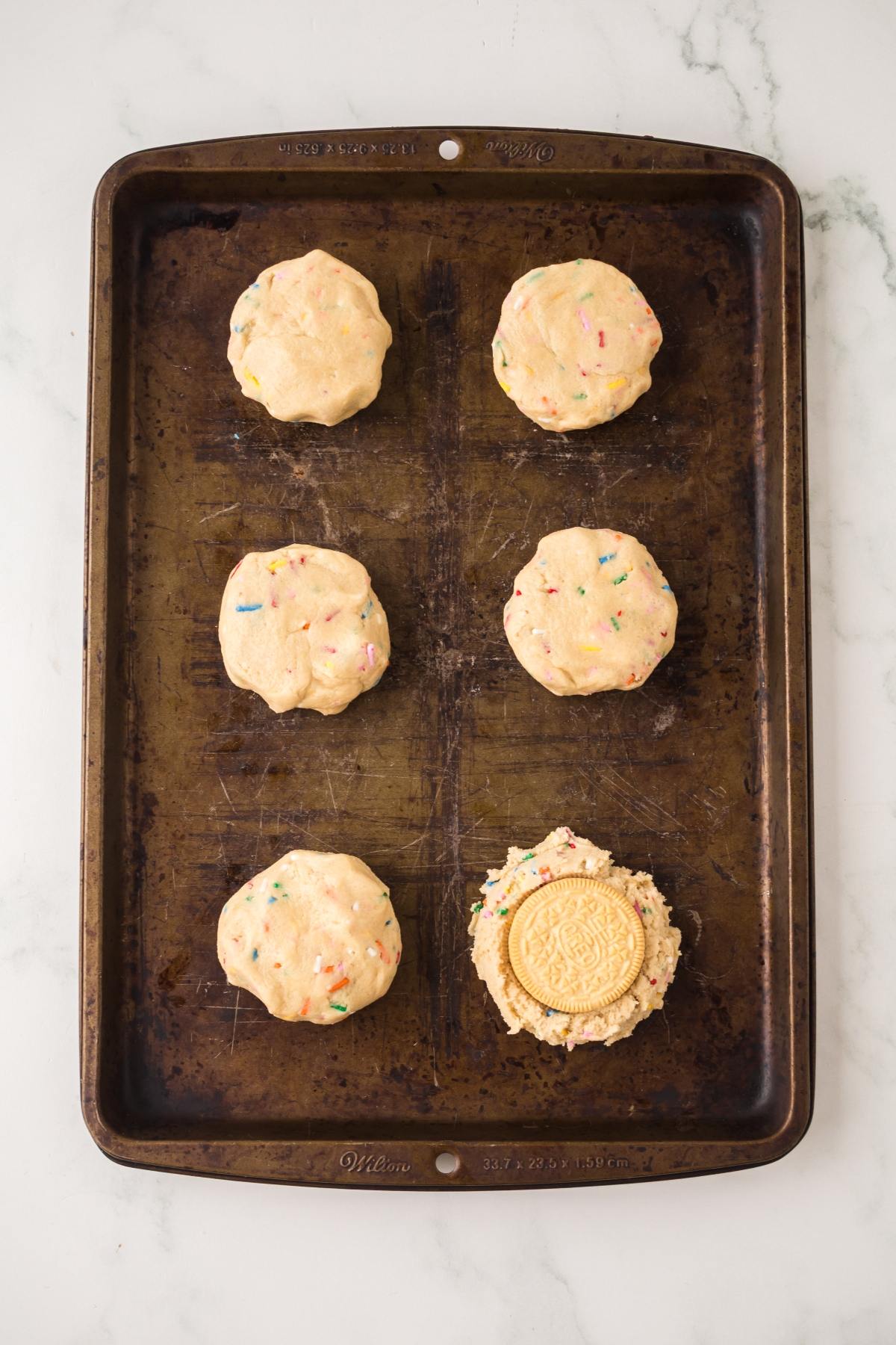 Six unbaked sprinkle cookie dough balls, one with a cookie inside, on a baking sheet.