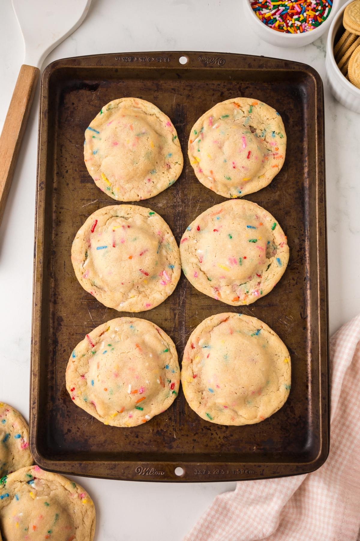Six large, round cookies with colorful sprinkles on a baking sheet, surrounded by baking tools.