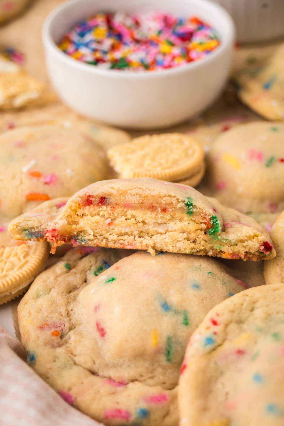 Funfetti cookies with colorful sprinkles, one broken in half, with Oreos and a bowl of sprinkles in the background.