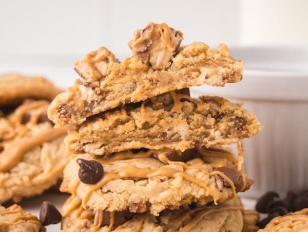 A stack of Peanut Butter Snickers Cookies drizzled with peanut butter, with a white ramekin in the background.