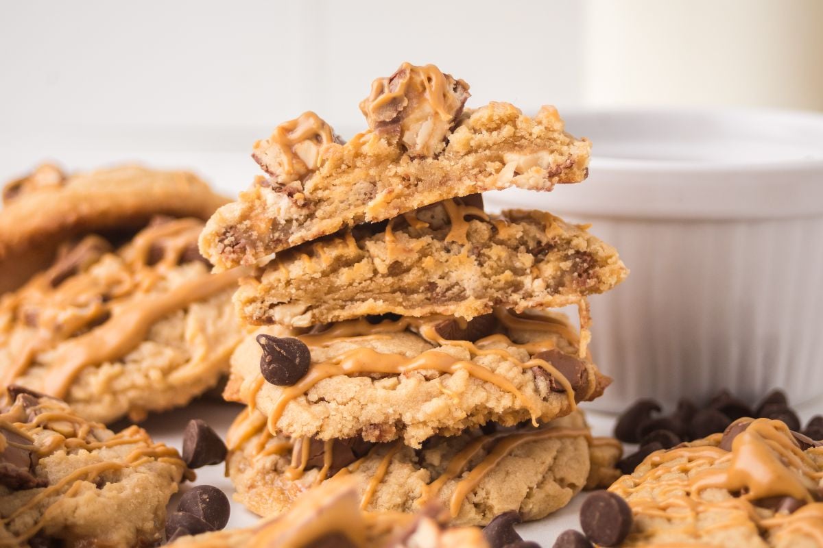 A stack of Peanut Butter Snickers Cookies drizzled with peanut butter, surrounded by chocolate chips.