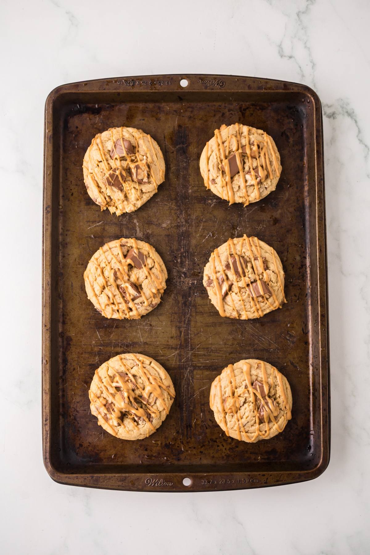 Six large cookies with drizzled topping on a baking tray, on a white marble surface.