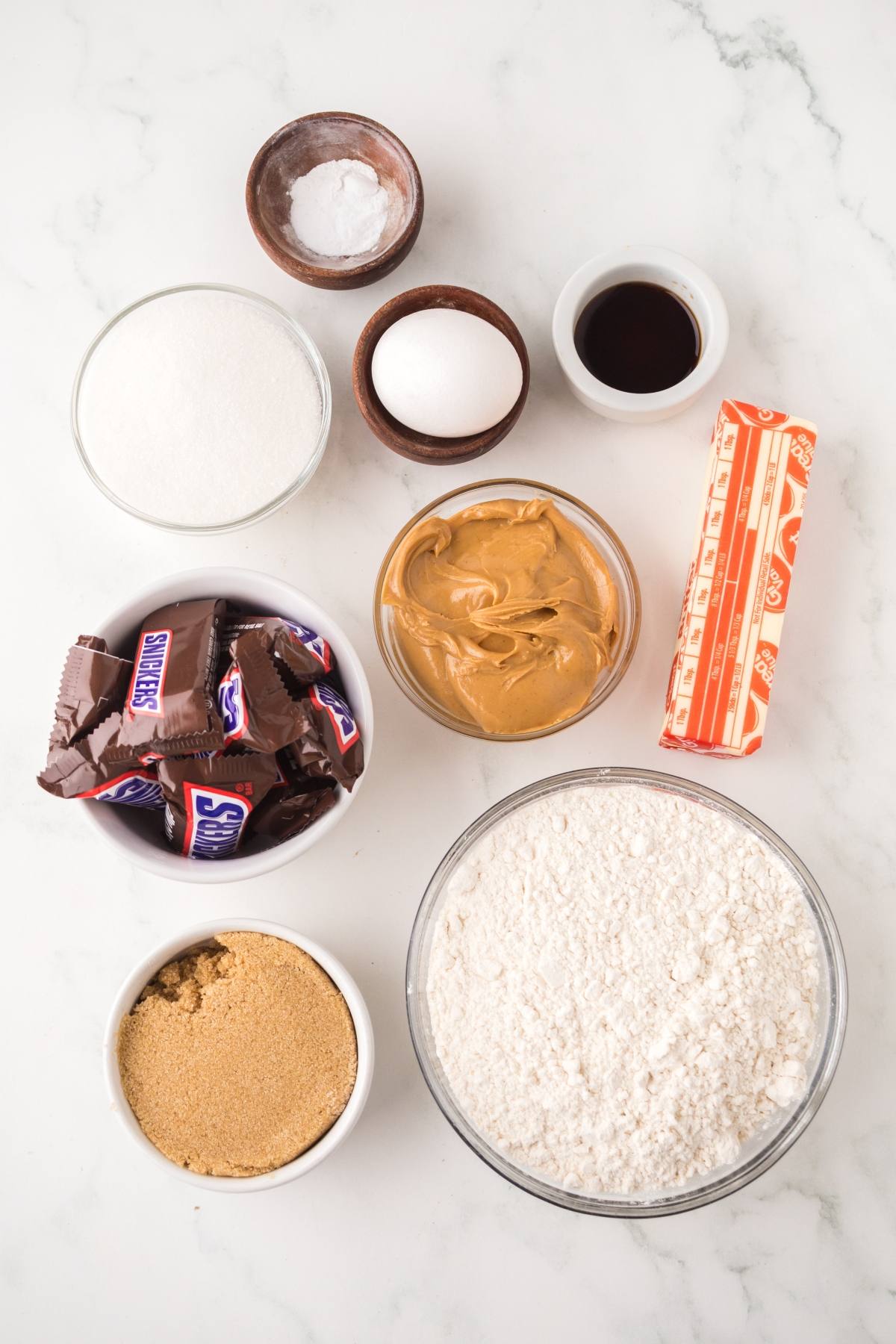 Baking ingredients on a white surface: flour, sugars, egg, butter, peanut butter, candy bars, vanilla, and baking soda.
