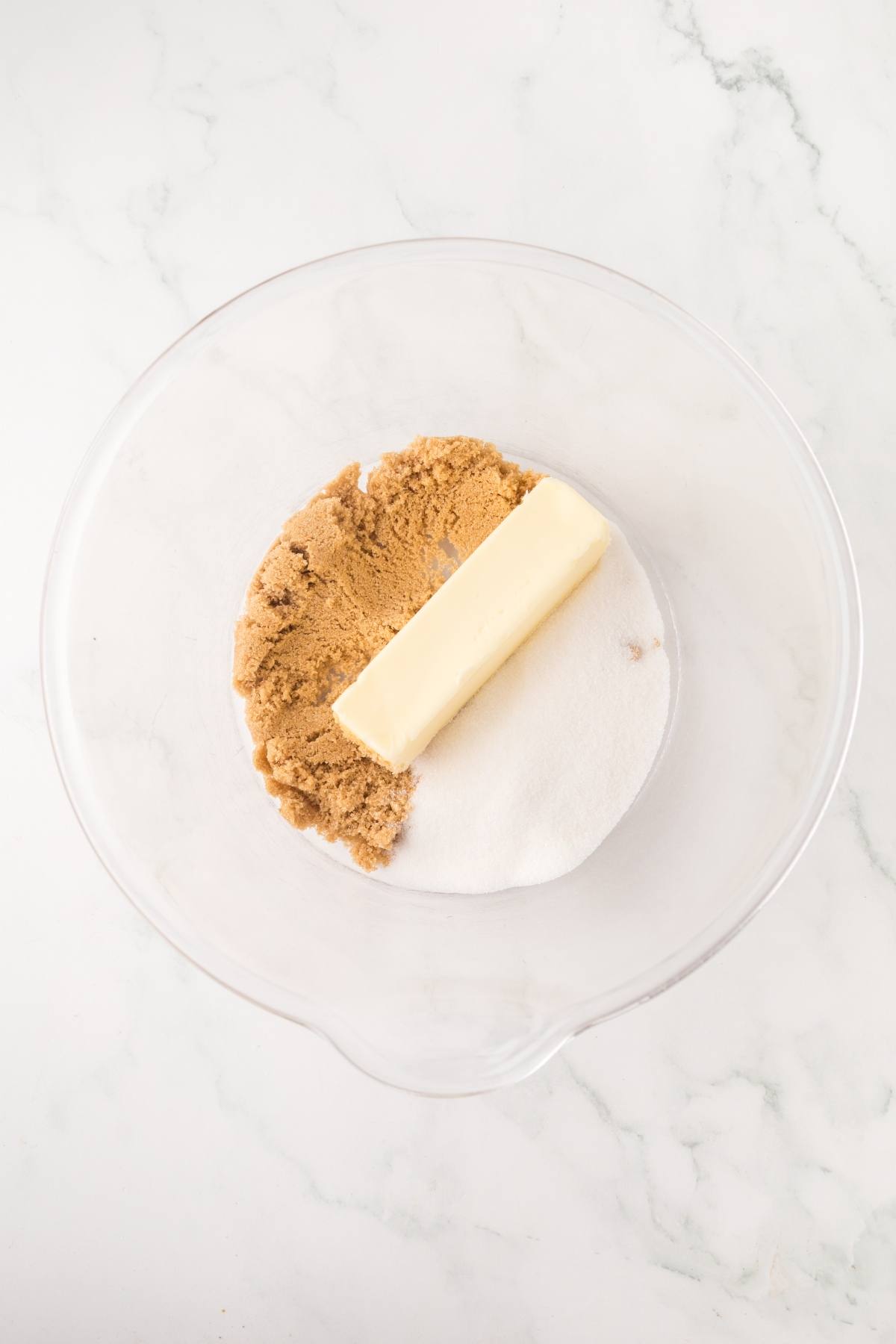 Clear bowl with white sugar, brown sugar, and a stick of butter on a white marble surface.