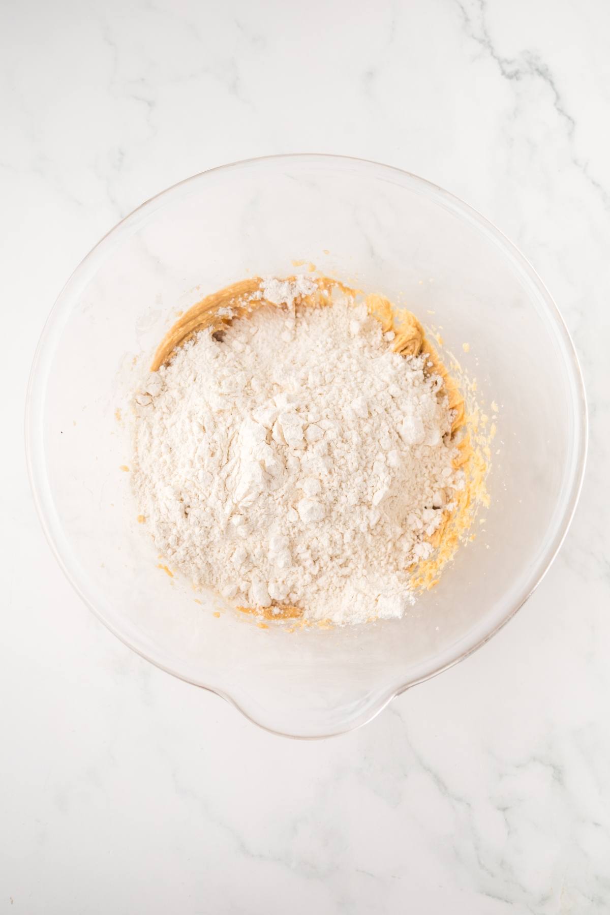 A glass mixing bowl with flour and dough mixture on a white marble surface.