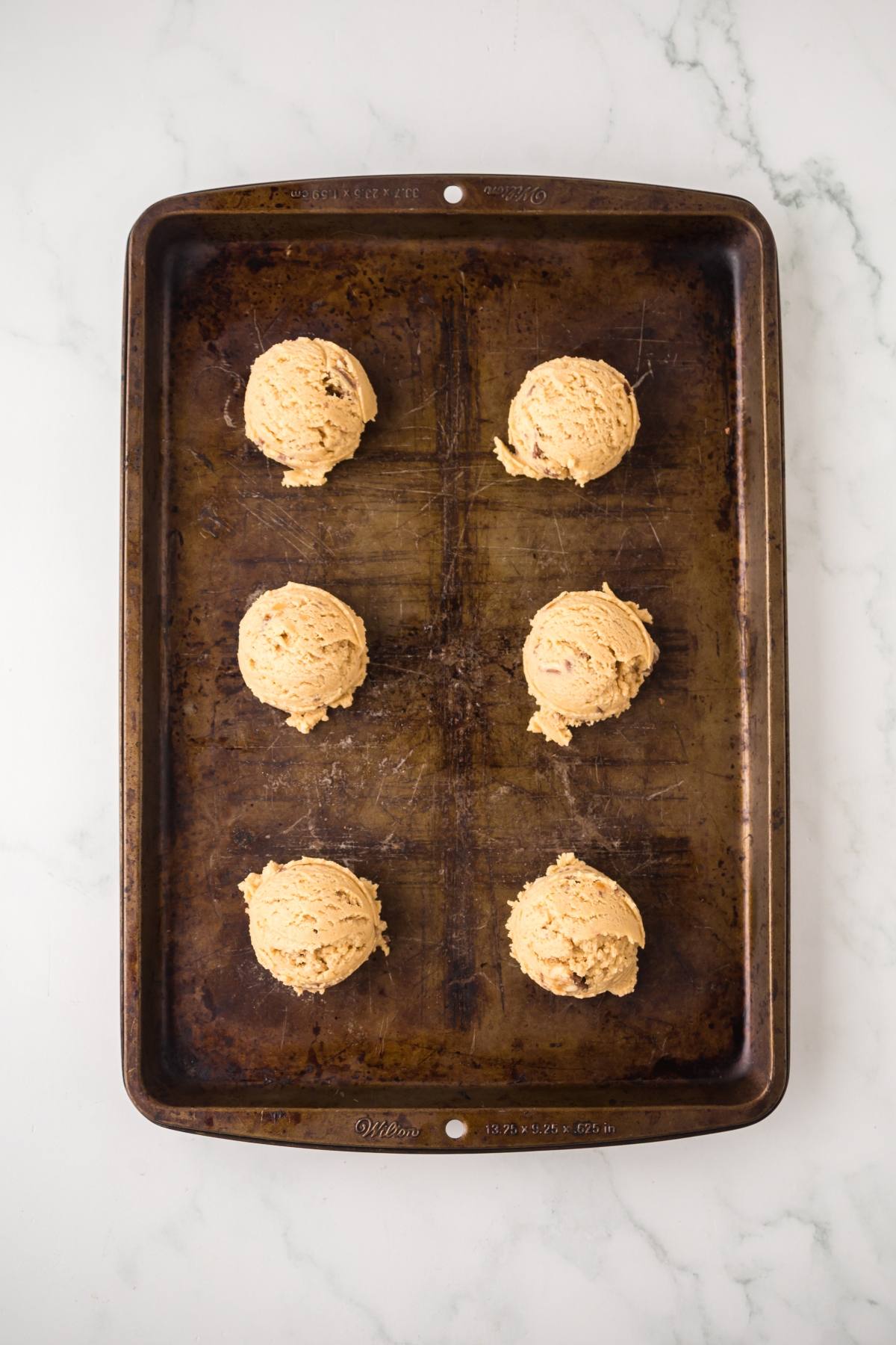 Six scoops of cookie dough spaced on a worn baking sheet, ready to be baked, on a marble surface.
