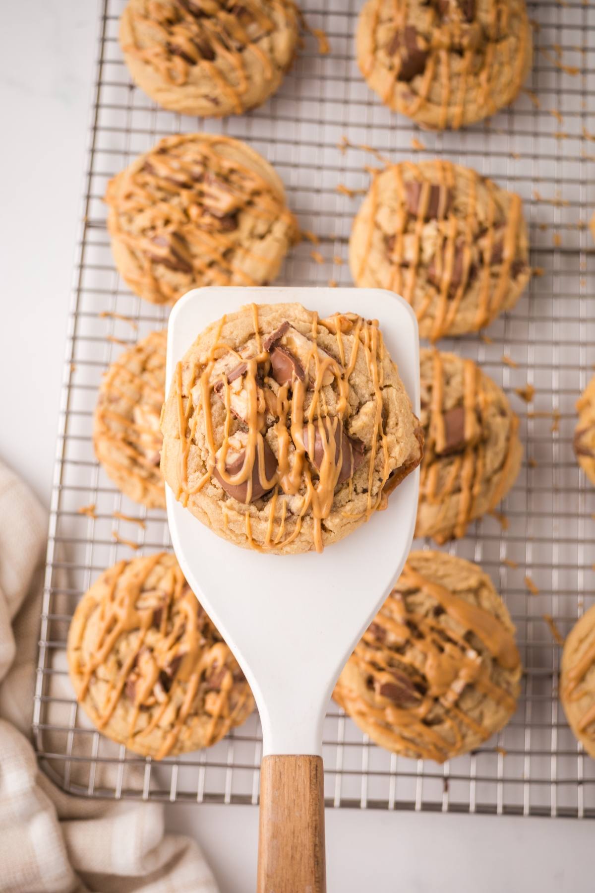 A spatula holds a peanut butter cookie with drizzle above a cooling rack of more cookies.