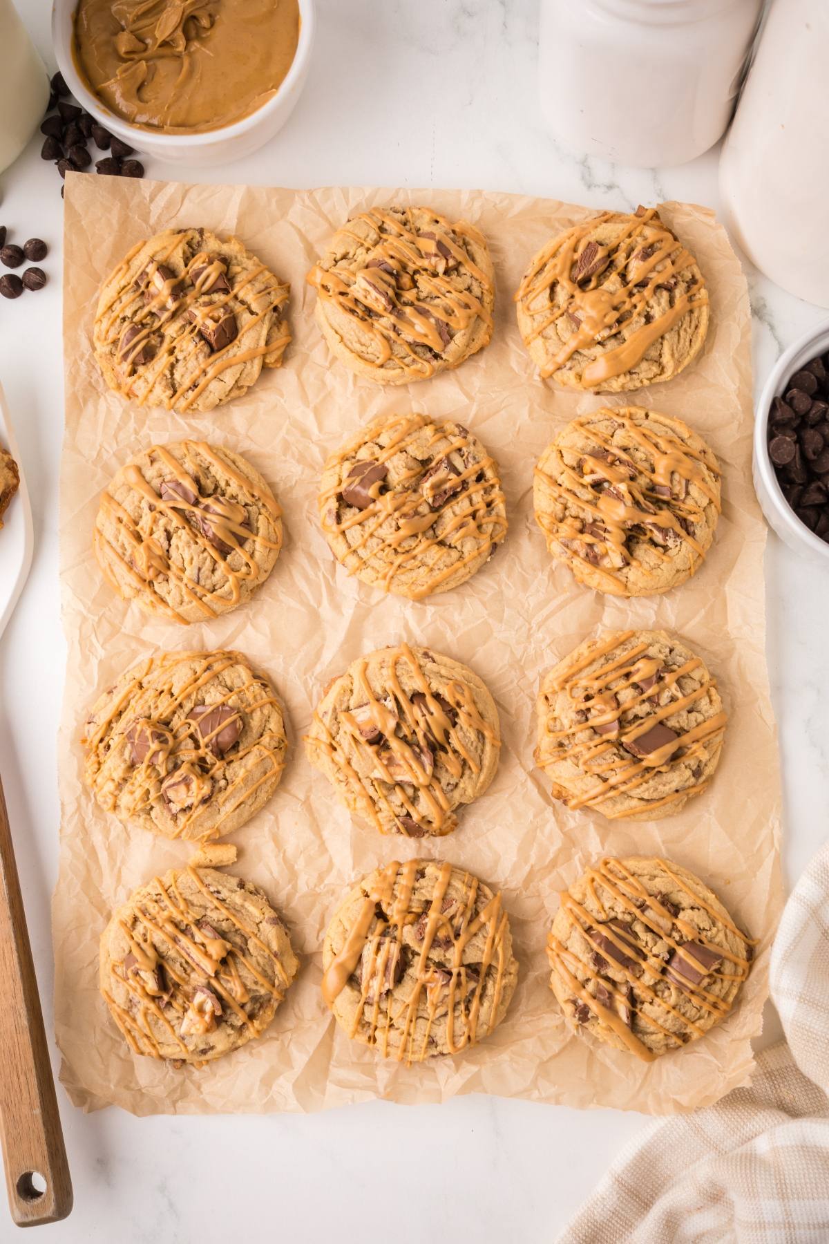 Twelve peanut butter cookies drizzled with peanut butter on parchment paper, surrounded by baking ingredients.